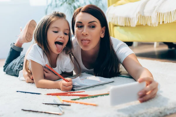 Mère et fille heureuses avec des langues qui sortent en prenant selfie tout en étant couchées sur le sol avec des crayons de couleur sur le premier plan flou — Photo de stock