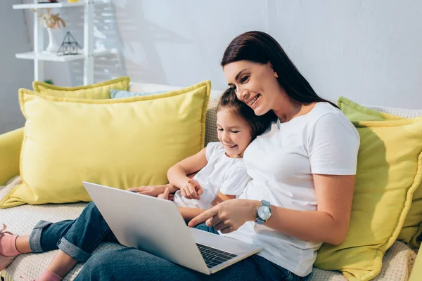 Feliz madre e hija riendo mientras señalan con los dedos a la computadora portátil en el sofá sobre un fondo borroso - foto de stock