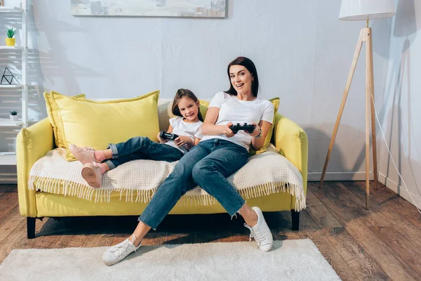 KYIV, UKRAINE - OCTOBER 19, 2020: Full length of cheerful mother and daughter looking at camera while holding joysticks on couch — Stock Photo