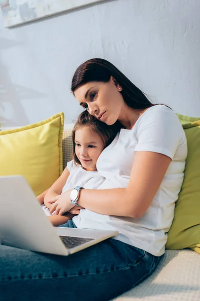 Madre che abbraccia sorridente figlia mentre guarda il laptop sul divano in primo piano sfocato — Foto stock