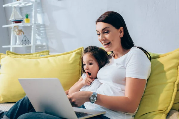 Emocionada hija y madre riendo mientras mira el portátil en el sofá sobre fondo borroso - foto de stock