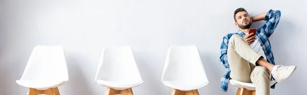 Young man using smartphone while sitting on chair in hall, banner — Stock Photo