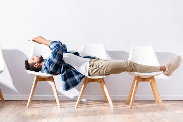 Young man using smartphone while lying on chairs in hall — Stock Photo