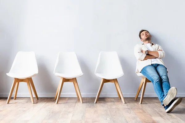 Hombre cansado sentado con los brazos cruzados cerca de sillas en el pasillo - foto de stock