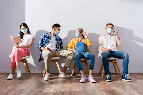 Multicultural people in medical masks talking and reading book on chairs in hall — Stock Photo