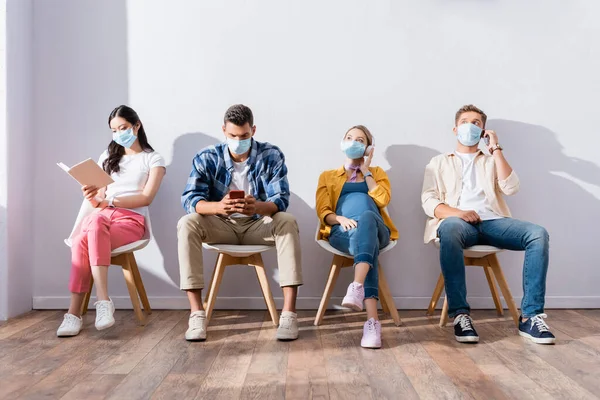 Pessoas multiculturais em máscaras médicas usando celulares, fones de ouvido e livro em fila — Fotografia de Stock