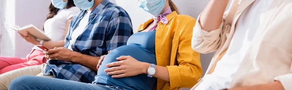 Обрізаний вид вагітної жінки в медичній масці, що сидить поруч з людьми в черзі на розмитому тлі, банер — стокове фото