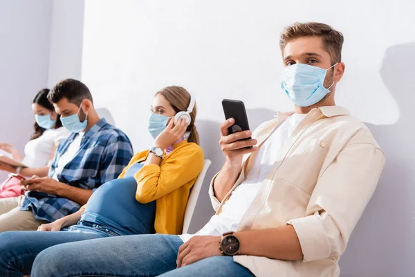 Man in medical mask holding smartphone near pregnant woman in queue — Stock Photo