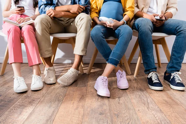 Cropped view of young people with smartphones and book sitting on chairs in hall — Stock Photo