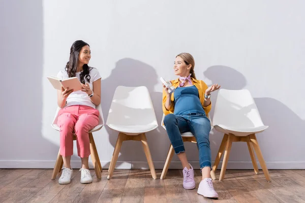 Smiling pregnant woman holding headphones near asian woman with book in queue — Stock Photo