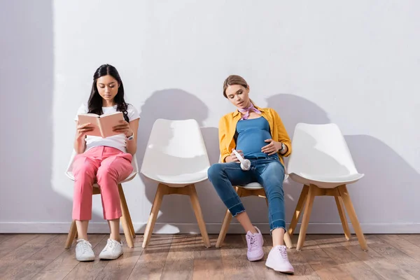 Pregnant woman with headphones sitting near asian woman reading book on chairs in hall — Stock Photo