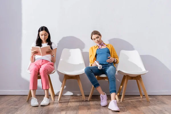 Embarazada mujer con auriculares sentado cerca asiático mujer leyendo libro en sillas en hall - foto de stock