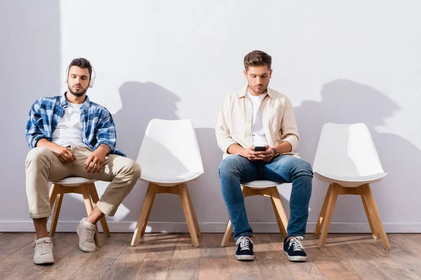 Hombres jóvenes usando auriculares y teléfonos inteligentes en sillas mientras esperan en la sala - foto de stock