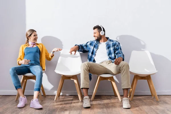 Homme souriant dans les écouteurs pointant vers la femme enceinte sur les chaises dans le hall — Photo de stock