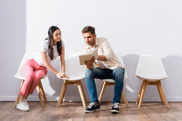 Hombre holding libro cerca asiático mujer en sillas en hall - foto de stock