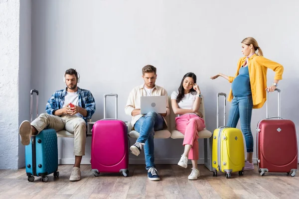 Femme enceinte pointant de la main près des personnes multiethniques avec des appareils et des valises à l'aéroport — Photo de stock