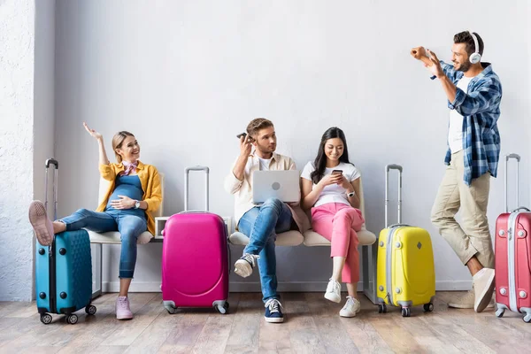 Donna incinta sorridente che saluta l'uomo in cuffia vicino a persone multietniche con dispositivi e valigie in aeroporto — Foto stock