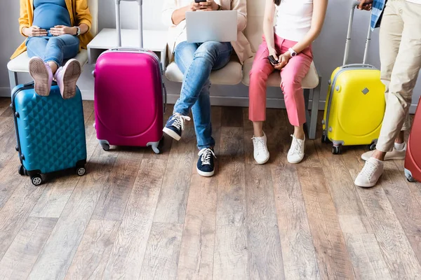 Vista recortada de la mujer embarazada sentada cerca de personas con dispositivos y maletas en el aeropuerto — Stock Photo