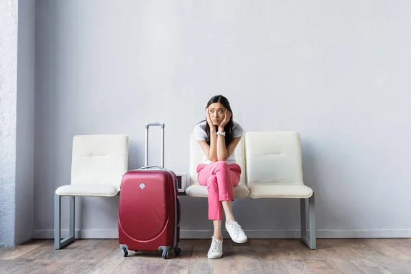 Уставшая азиатская женщина смотрит в сторону рядом с чемоданом во время ожидания в аэропорту — стоковое фото