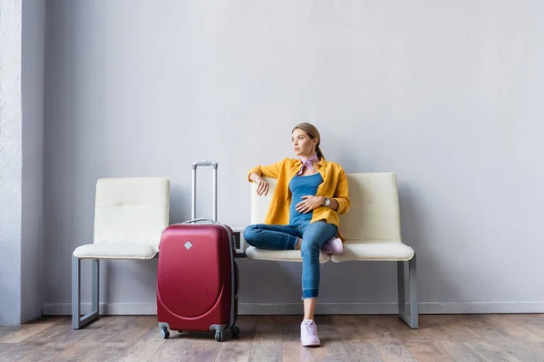 Mujer embarazada sentada cerca de la maleta mientras espera en el aeropuerto - foto de stock