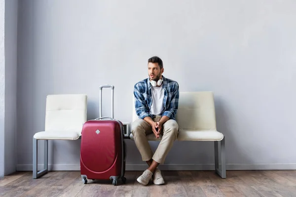 Hombre con auriculares mirando hacia otro lado cerca de la maleta en el aeropuerto - foto de stock