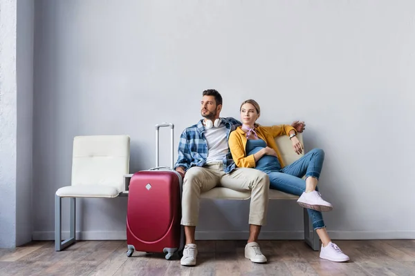 Donna incinta seduta vicino a marito e valigia in aeroporto — Foto stock