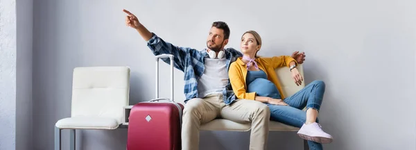 Чоловік вказує пальцем біля вагітної дружини і валізи в аеропорту, банер — стокове фото
