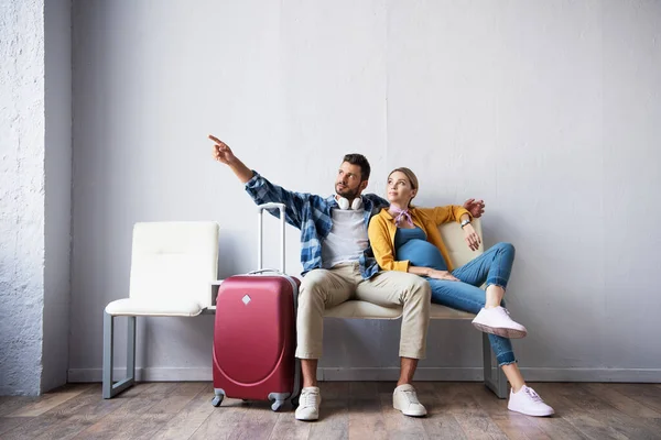 Hombre señalando con el dedo cerca de la esposa embarazada y el equipaje en la sala del aeropuerto - foto de stock