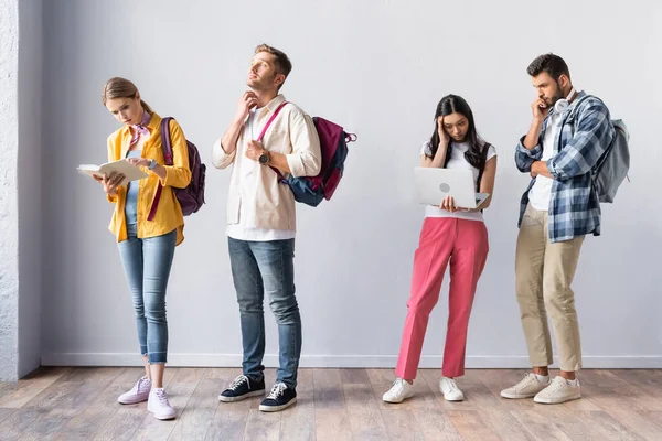 Estudiantes multiétnicos con libro y portátil de pie en la sala durante el examen - foto de stock