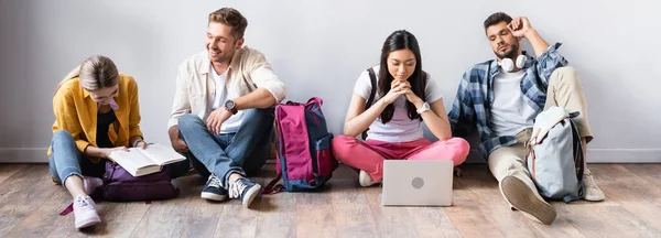 Estudiantes multiétnicos con portátil, libro y mochilas sentados en el suelo, pancarta — Stock Photo