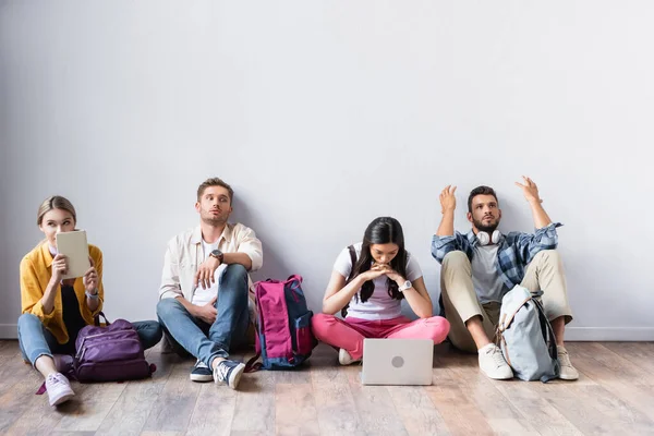 Багатоетнічні студенти з рюкзаками, ноутбуком і книгою чекають на підлозі в залі — стокове фото