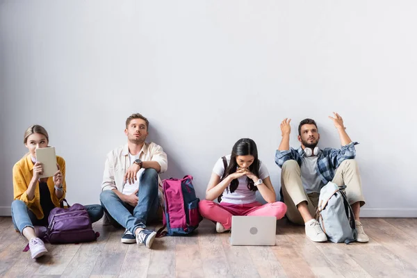 Estudiantes multiétnicos con mochilas, laptop y libro esperando en el piso en la sala - foto de stock