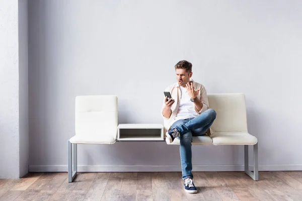 Hombre agresivo usando teléfono inteligente mientras está sentado en la silla en el pasillo - foto de stock