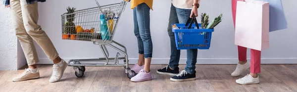 Cropped view of people holding shopping bags, cart and basket with food in queue, banner — Stock Photo