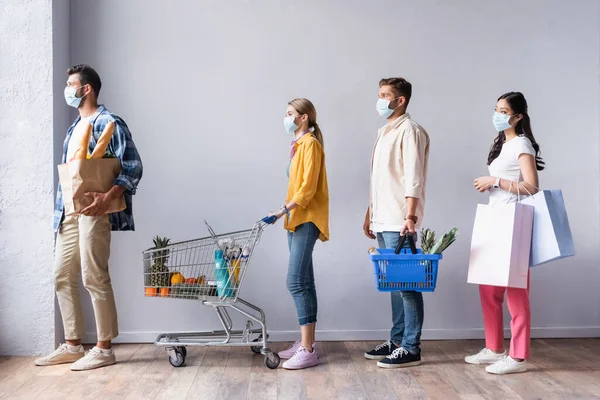 Gente multicultural en máscaras médicas sosteniendo carrito de la compra, cesta con comida y bolsas en cola en el mercado - foto de stock