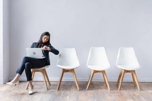 Mujer de negocios asiática con portátil mirando reloj de pulsera mientras espera en la sala - foto de stock