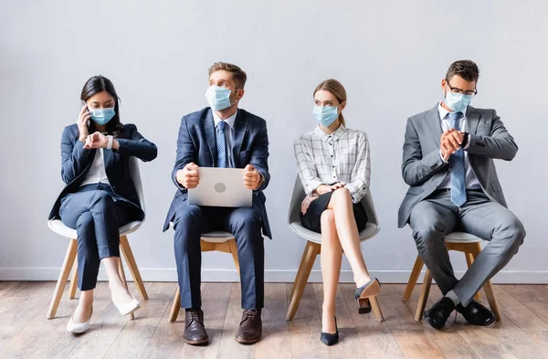 Gente multicultural en máscaras médicas con portátil y smartphone esperando en sillas en el pasillo — Stock Photo
