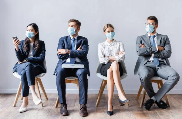 Gente de negocios multicultural en máscaras médicas con dispositivos esperando entrevista de trabajo en la sala - foto de stock