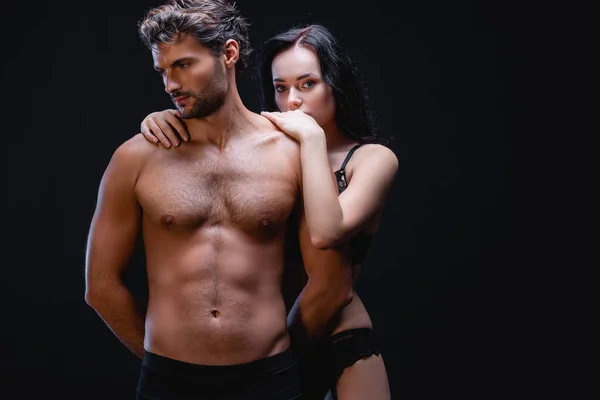 Femme brune sensuelle embrassant les épaules de l'homme avec le torse musculaire tout en regardant la caméra isolée sur noir — Photo de stock