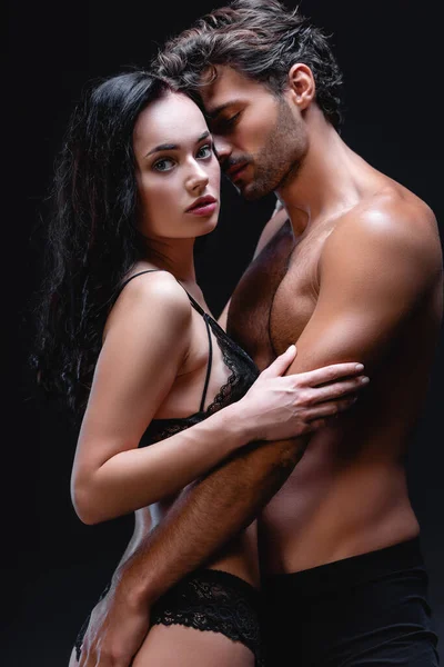 Сексуальний без сорочки чоловік обіймає чуттєву брюнетку жінку, дивлячись на камеру ізольовано на чорному — стокове фото