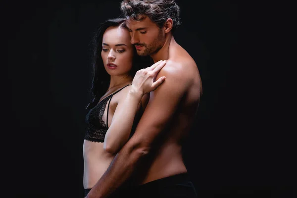 Joven sin camisa hombre abrazando sexy mujer en encaje sujetador aislado en negro - foto de stock