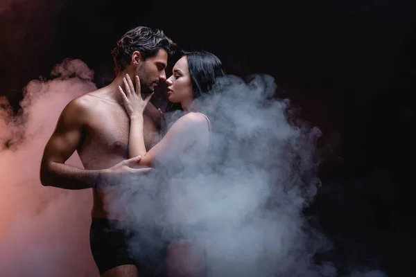 Sensual woman touching sexy shirtless man while standing face to face on dark background with smoke — Stock Photo