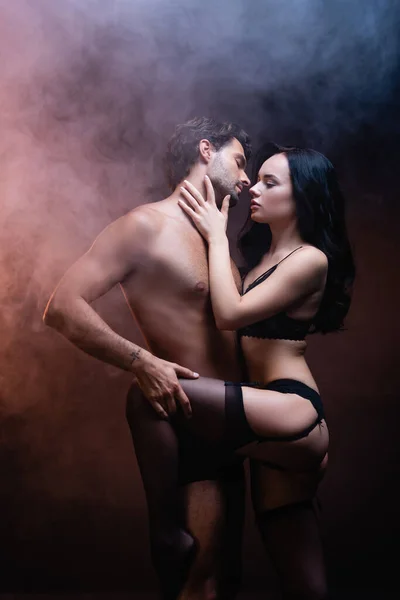 Femme sexy en lingerie noire et bas touchant le cou de l'homme torse nu sur fond sombre avec de la fumée — Photo de stock