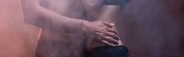 Vista ritagliata di uomo tatuato senza maglietta che abbraccia la donna su sfondo scuro con fumo, banner — Foto stock