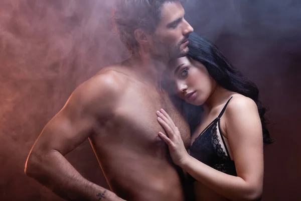 Sexy woman leaning on chest of shirtless muscular man while looking at camera on dark background with smoke — Stock Photo