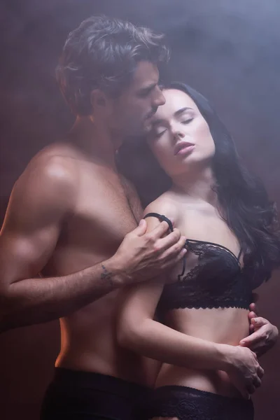 Hombre musculoso sin camisa abrazando a mujer seductora en sujetador de encaje negro sobre fondo oscuro con humo - foto de stock