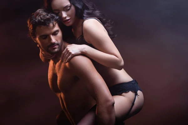 Sexy shirtless man piggybacking seductive woman in black underwear on dark background — Stock Photo