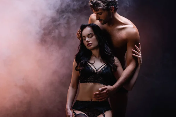 Sexy shirtless man touching seductive woman in black lace underwear on dark background with smoke — Stock Photo