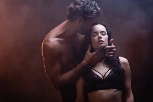 Hemdloser muskulöser Mann umarmt Hals der verführerischen Frau in schwarzem Spitzen-BH auf dunklem Hintergrund mit Rauch — Stockfoto