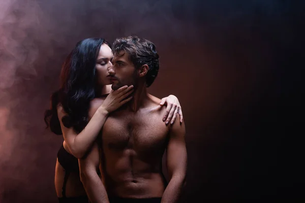 Соблазнительная женщина обнимает и целует сексуального мужчину без рубашки на темном фоне с дымом — стоковое фото
